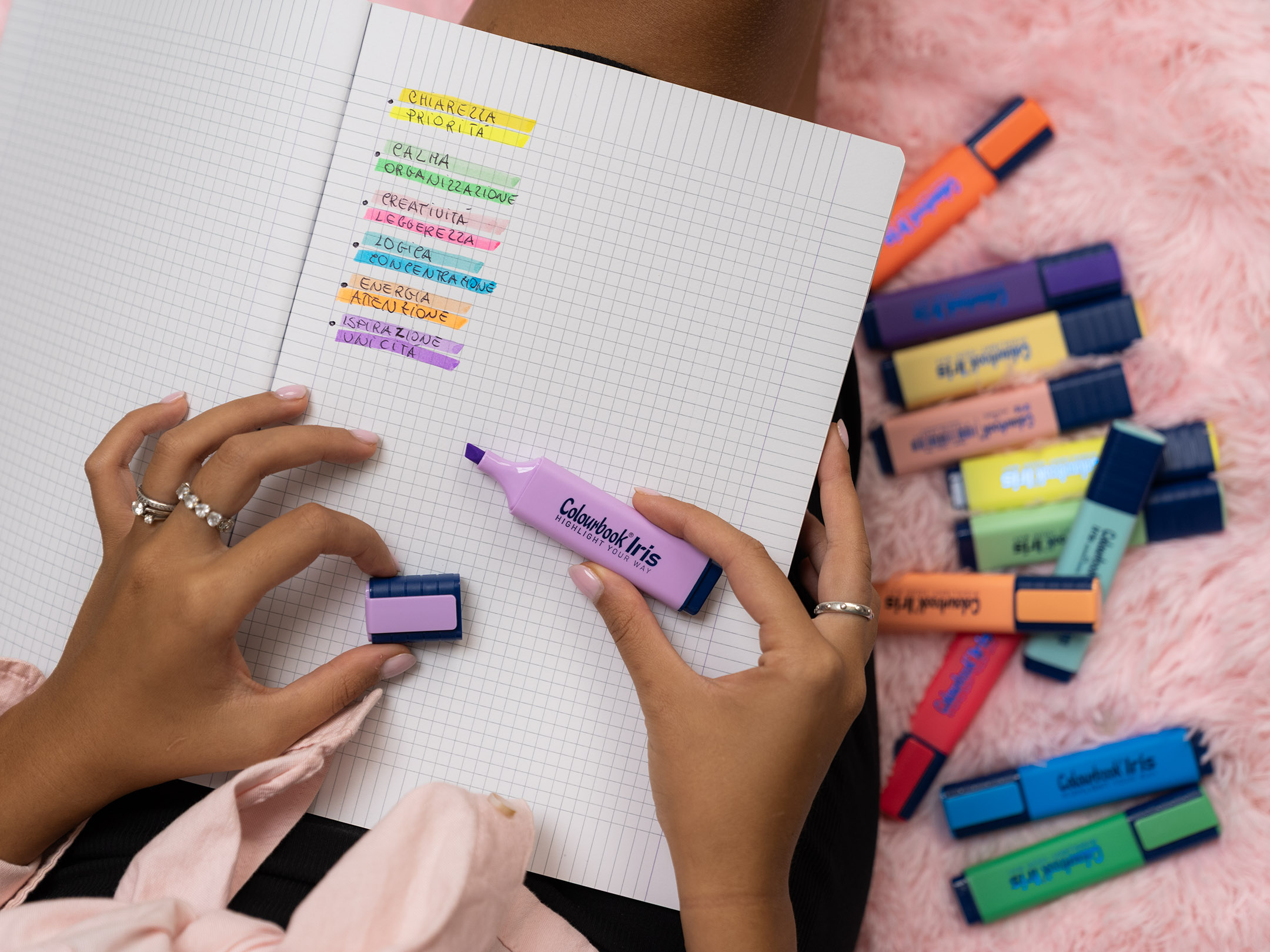 la tecnica del color code In questa immagine vediamo una mano che utilizza un evidenziatore lilla Colourbook Iris su un quaderno a quadretti. Le righe sono organizzate con diversi colori, ognuno associato a una parola chiave come chiarezza, priorità, creatività, energia o ispirazione. Attorno al quaderno sono sparsi altri evidenziatori Colourbook in vivaci tonalità pastello e fluo. L’insieme mette in risalto l’uso del color code: ogni colore diventa un segnale visivo che aiuta a distinguere concetti, organizzare idee e dare più energia allo studio e al lavoro.