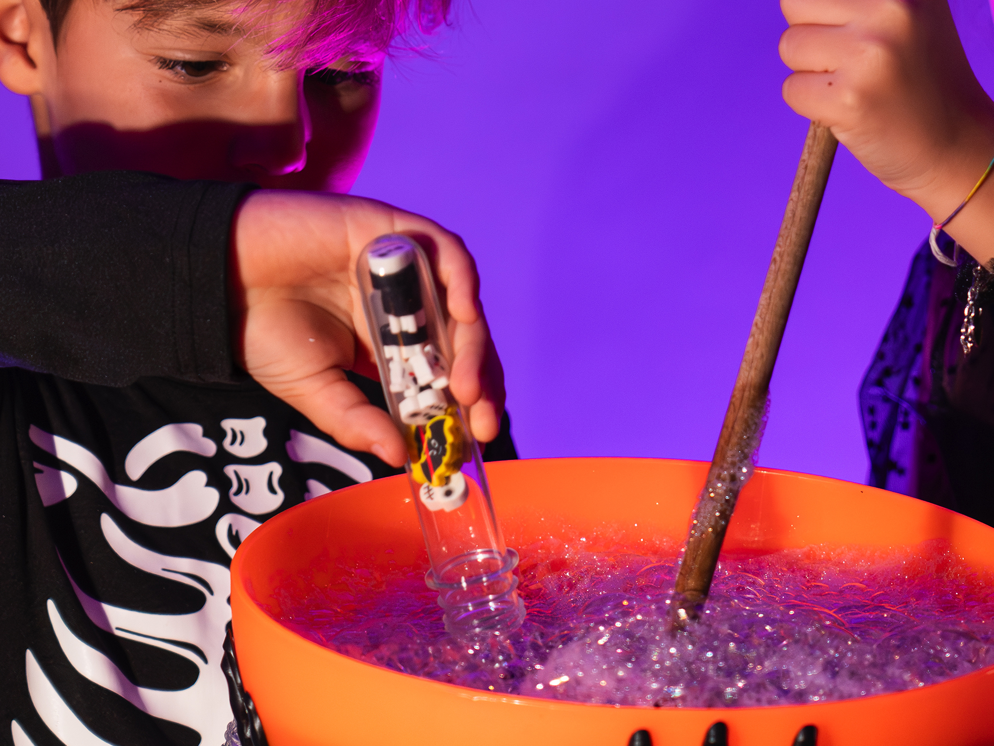 bambini che giocano Un bambino vestito da scheletro gioca con una zucca arancione piena di bolle scintillanti, mescolandole con un bastoncino come in un incantesimo. Nella mano tiene una provetta Colourbook con piccole decorazioni a tema Halloween. La scena, illuminata da luce viola e calda, trasmette un senso di magia e divertimento tipico della notte delle streghe.